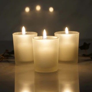 Three lit white candles in glass holders on a reflective surface with shadows and soft lighting.