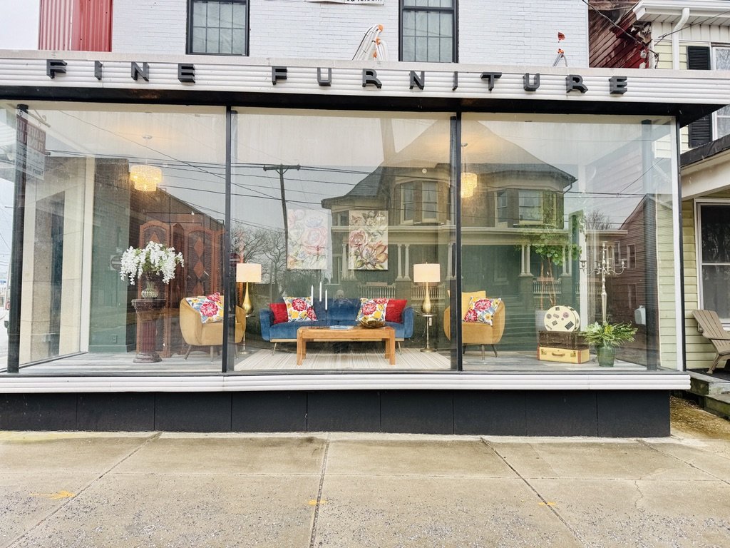 Storefront window display of a furniture store featuring a living room setup with a blue sofa, beige armchairs, colorful cushions, a wooden coffee table, floor lamps, and decorative flowers.