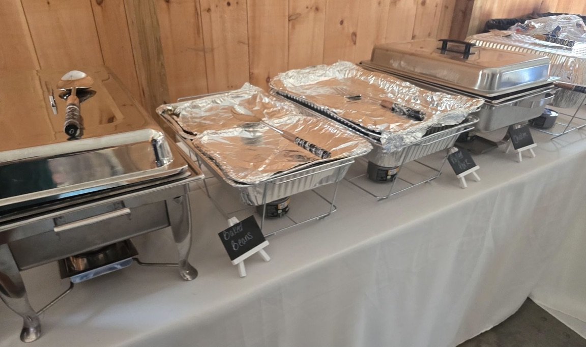 Buffet setup with chafing dishes and covered trays containing food, labels indicating contents such as baked beans.