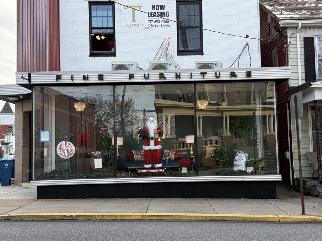 Storefront with a Christmas display featuring Santa Claus, holiday decorations, and a 'Merry Christmas' sign in a furniture store window.