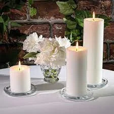 Three lit white pillar candles on glass holders with a small glass vase of white flowers in front of a brick wall and green foliage background.