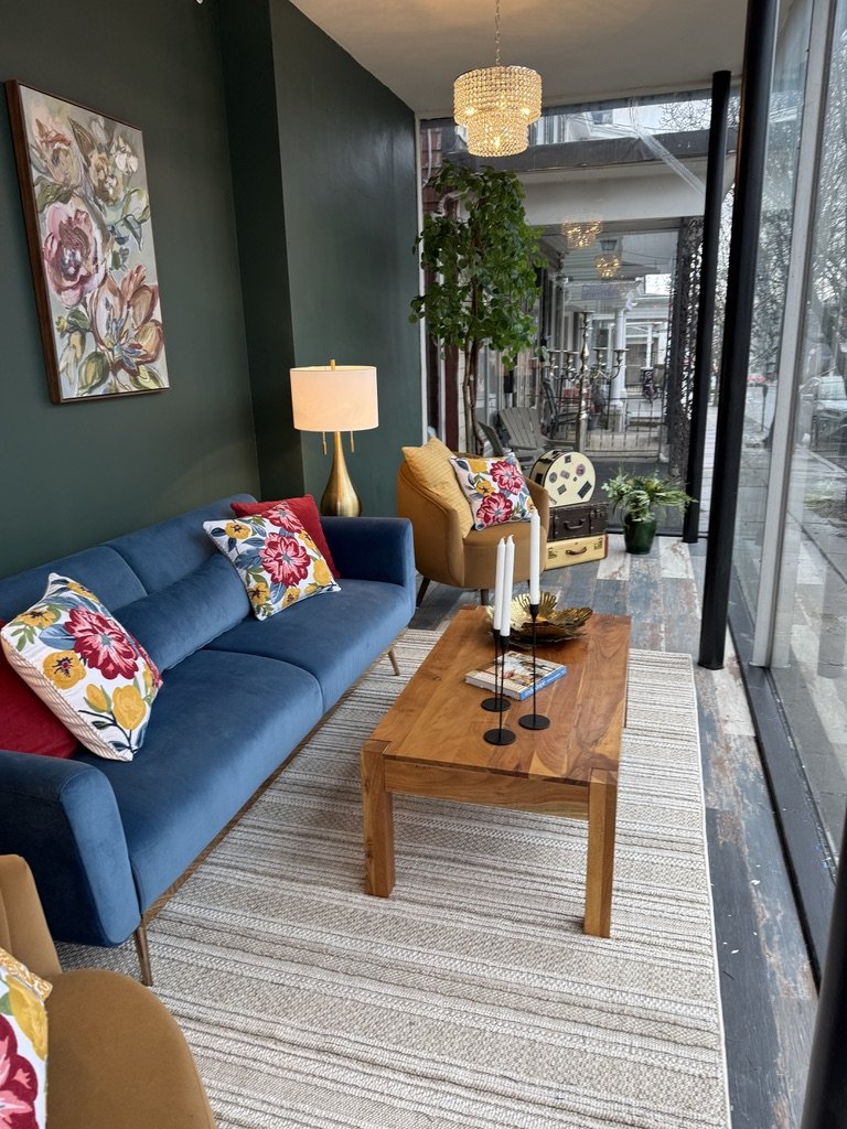 Modern living room with blue sofa, floral cushions, and wooden coffee table. Features a chandelier, large window, green wall art, and indoor plant.