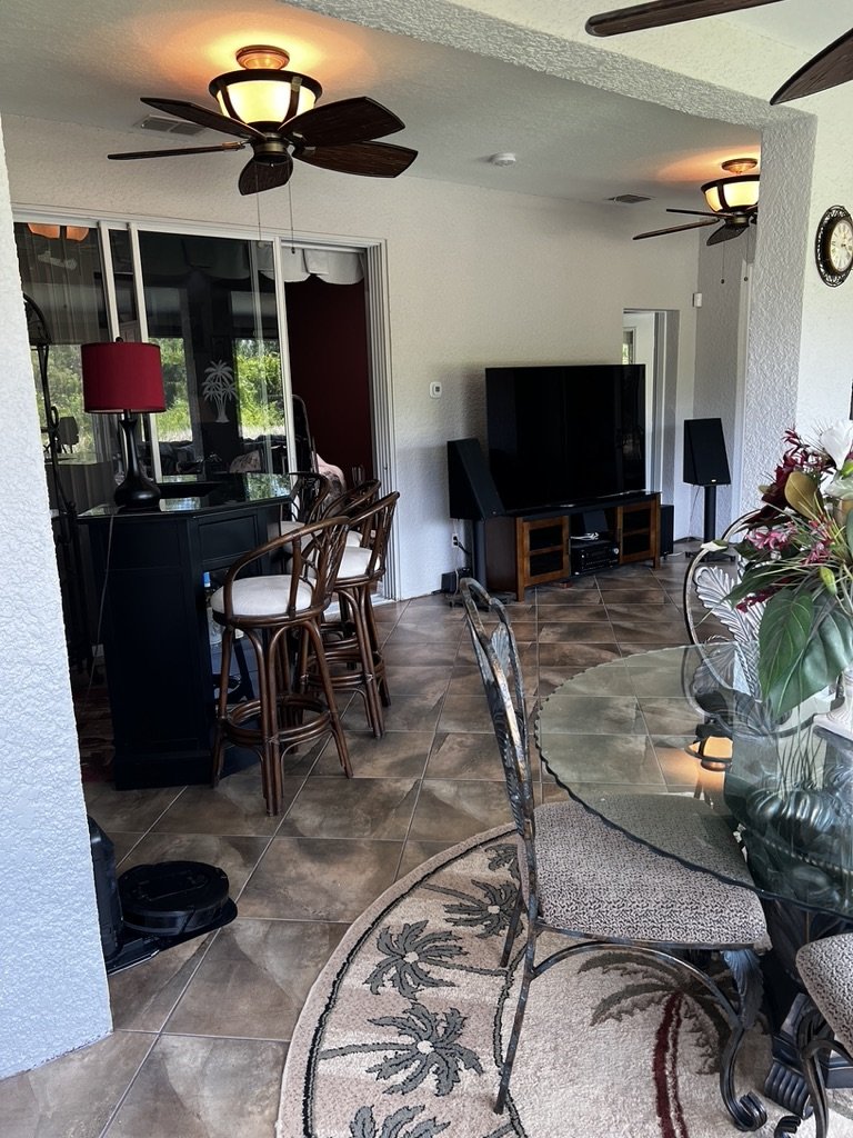 Indoor seating area with tiled floor, ceiling fans, bar stools, a glass-top table, chairs, and a large TV. Decor includes a round rug with palm tree design and a floral arrangement.
