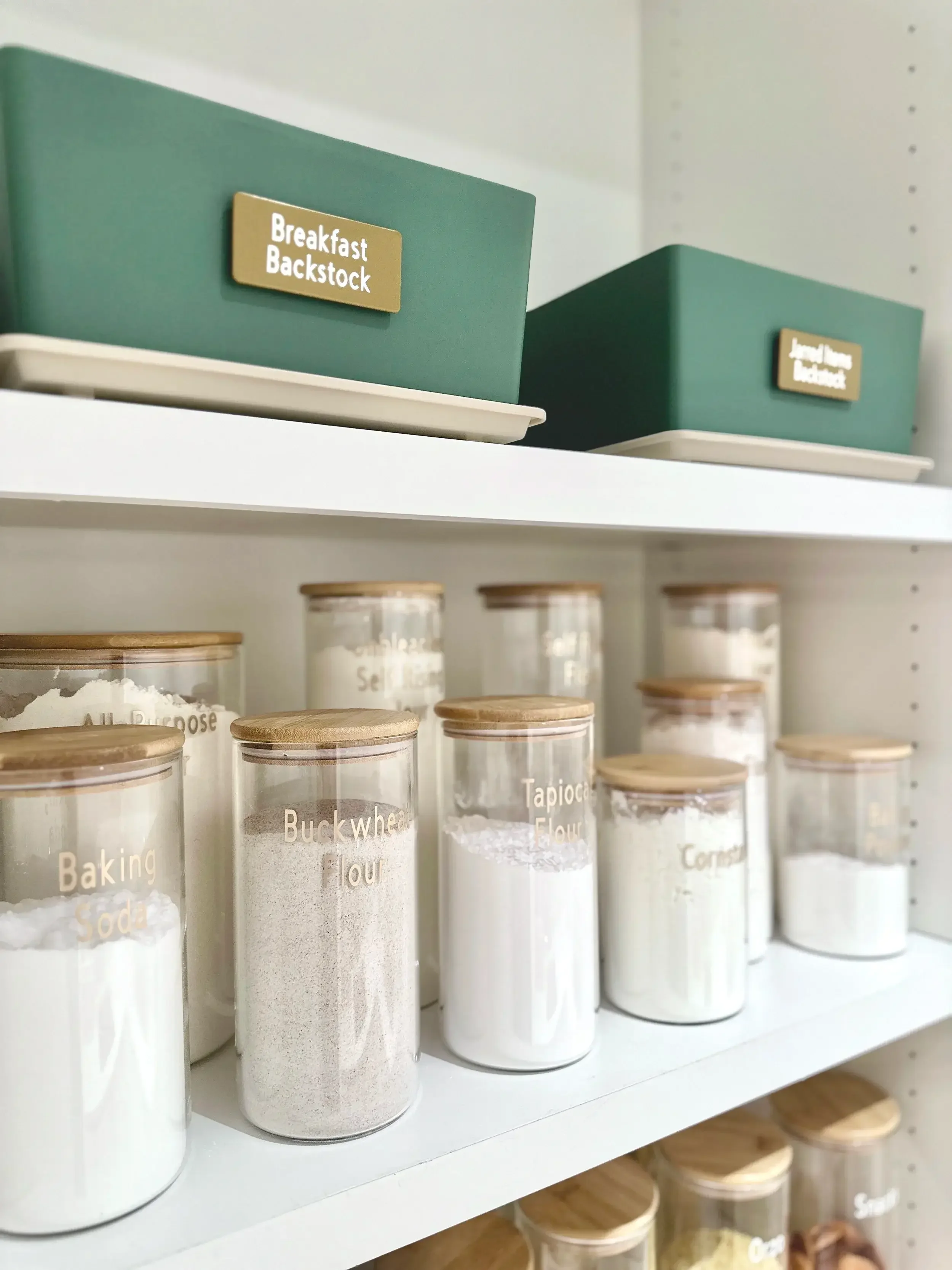 Shelf of clear glass jars with wooden lids labeled with baking ingredients, including flour, tapioca flour, cornmeal, and baking soda, with two green storage boxes labeled 'Breakfast Backstock' on top.