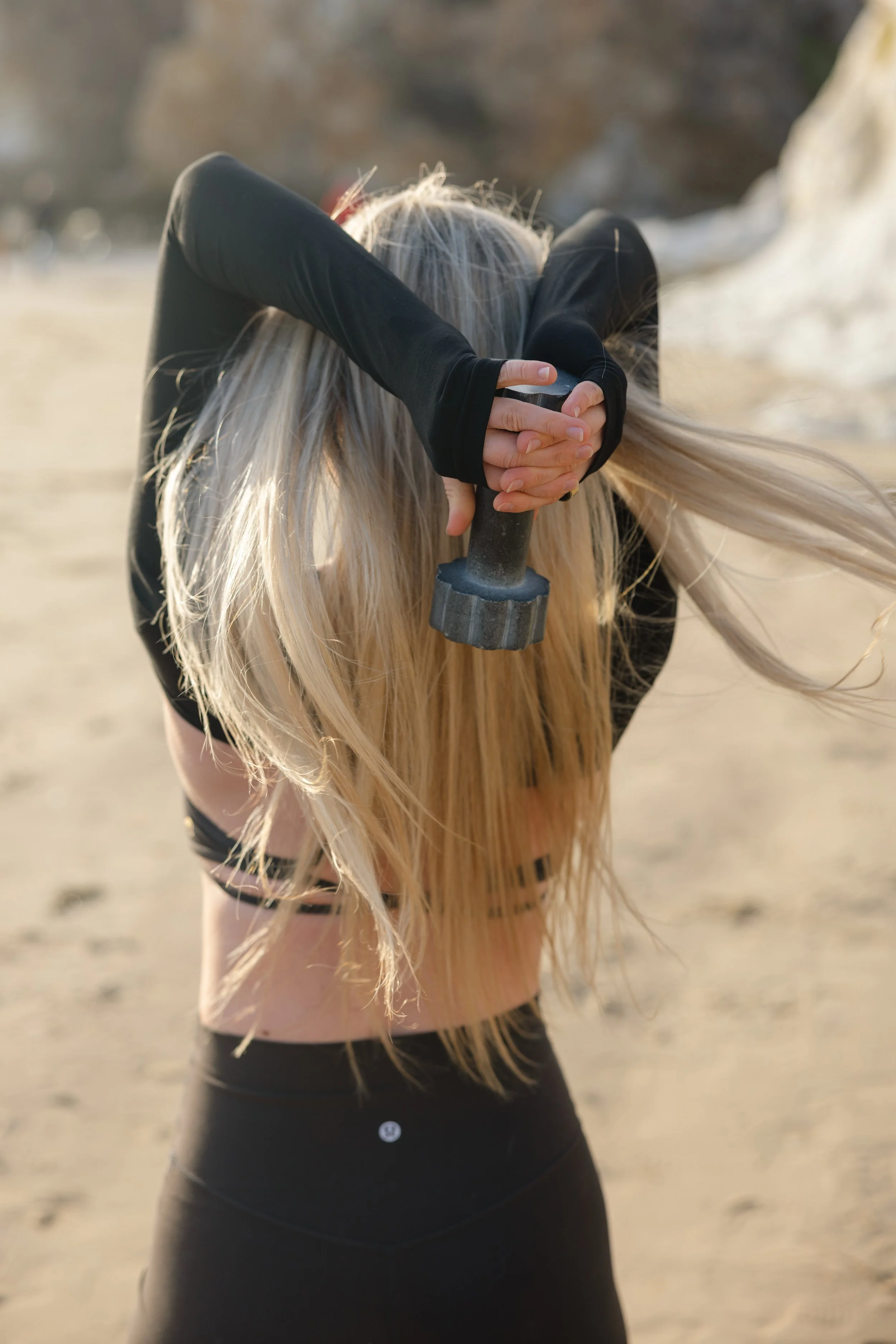 A woman with long blonde hair, wearing black athletic clothing, stretching her arms behind her head while holding a dumbbell, on a sandy beach with rocks and cliffs in the background.