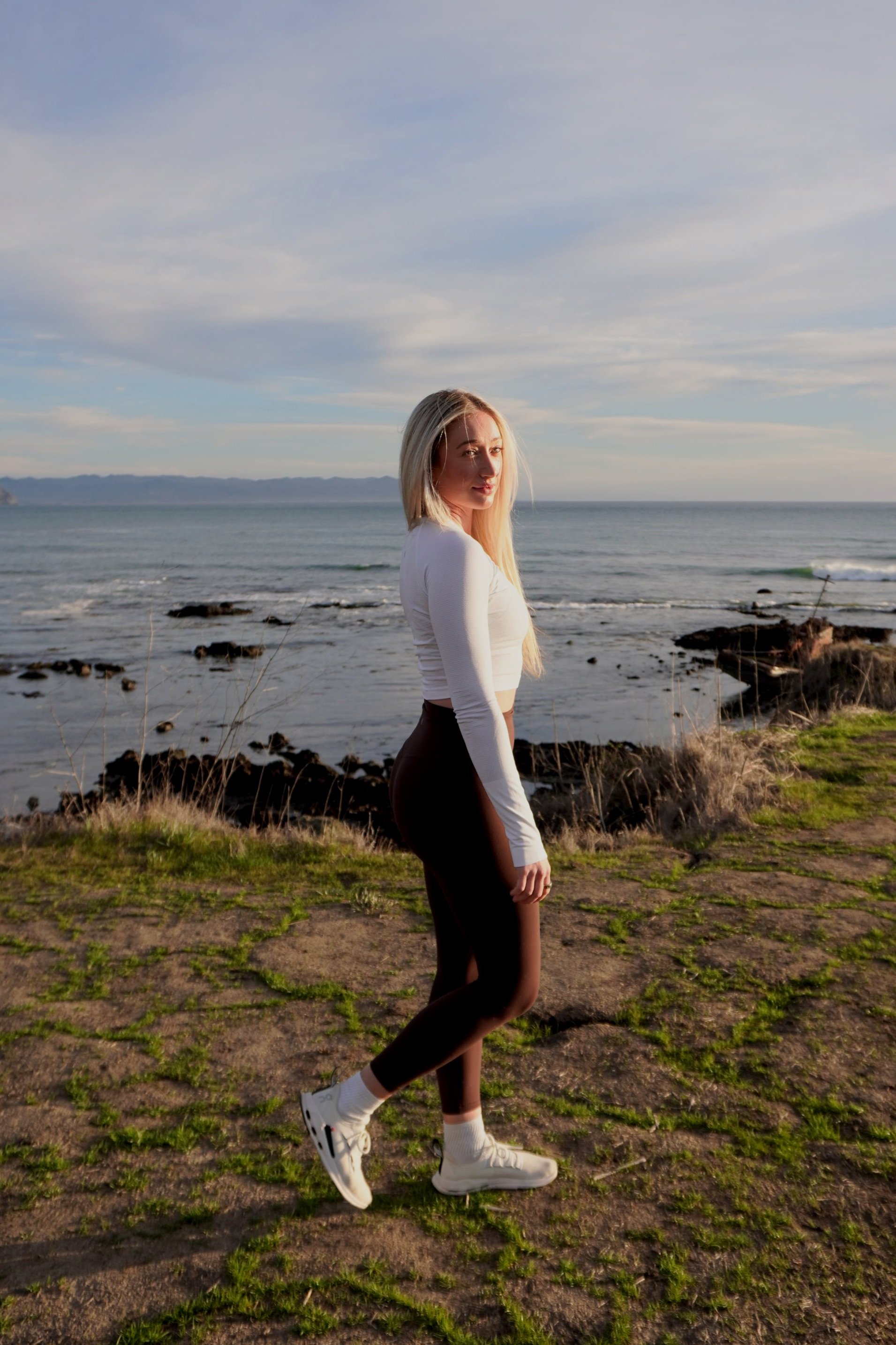 A person with long blonde hair in a white top and black leggings standing on a grassy area by the ocean during sunset.