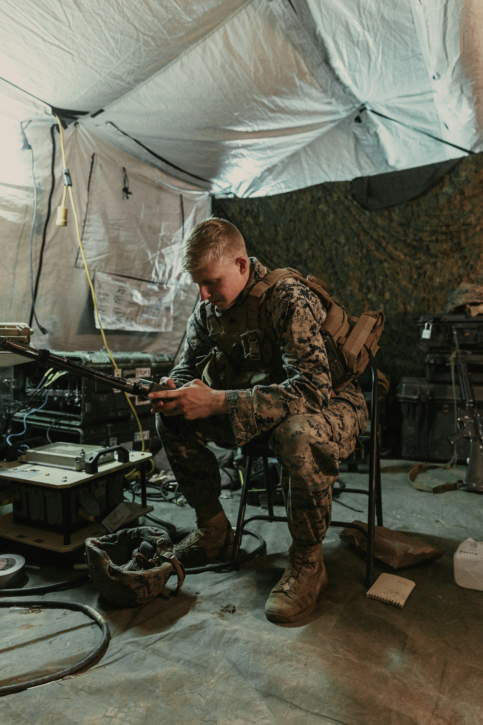 A soldier in camouflage uniform and gear sitting on a chair in a tent, working with electronic equipment, with various wires, tools, and personal items around him.