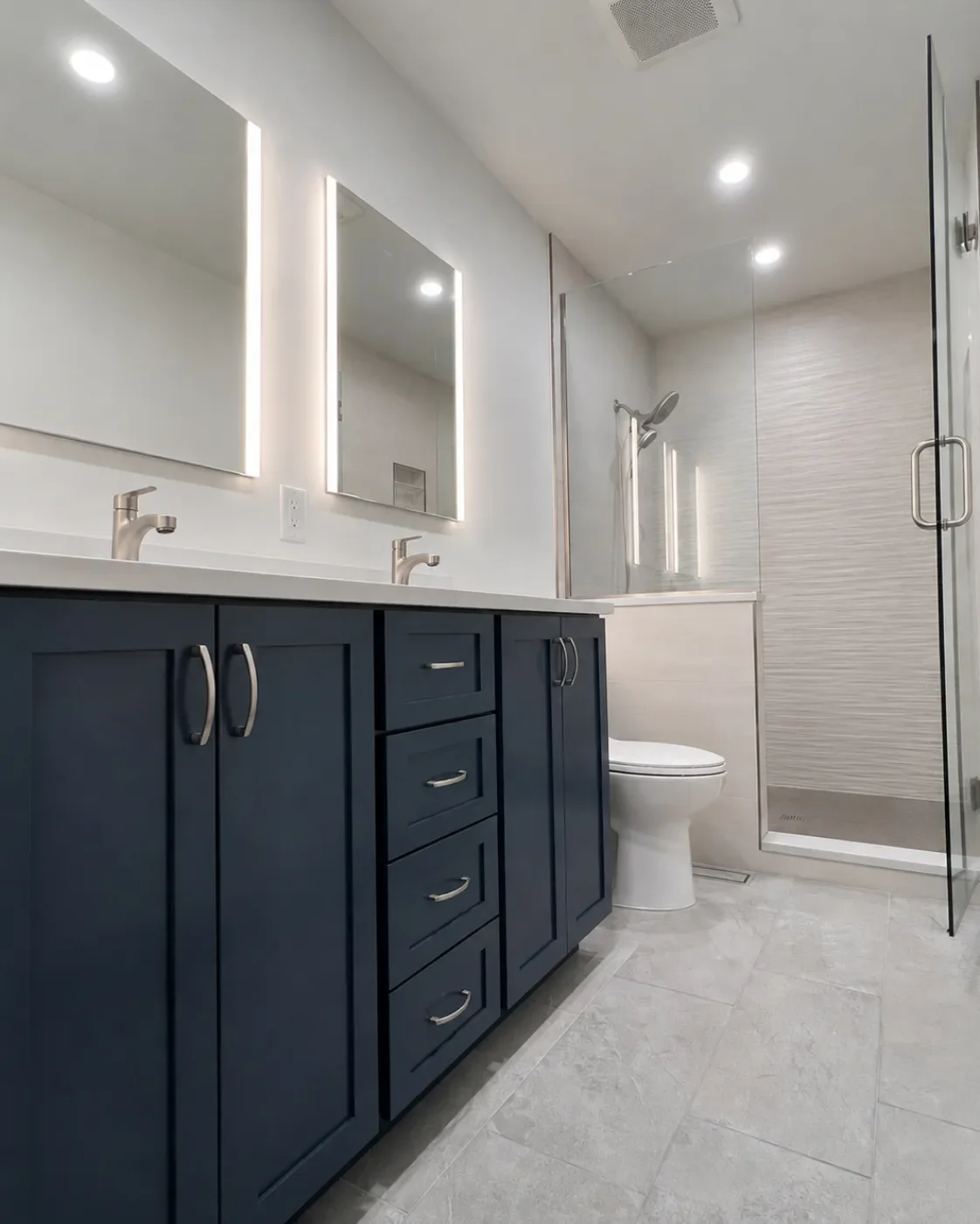 Contemporary bathroom remodel in Table Mesa, Boulder, CO featuring dark blue custom double vanity with recessed cabinet LED mirrors, frameless glass walk-in shower with textured accent wall, and gray stone tile flooring