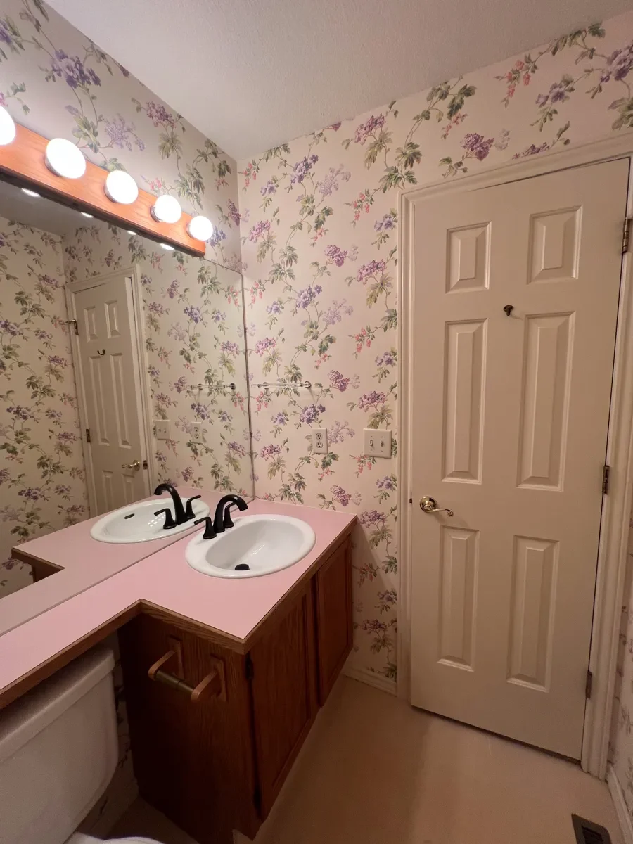 Gunbarrel, CO guest bathroom before renovation featuring outdated wooden vanity with pink laminate countertops, linoleum flooring, and floral wallpaper