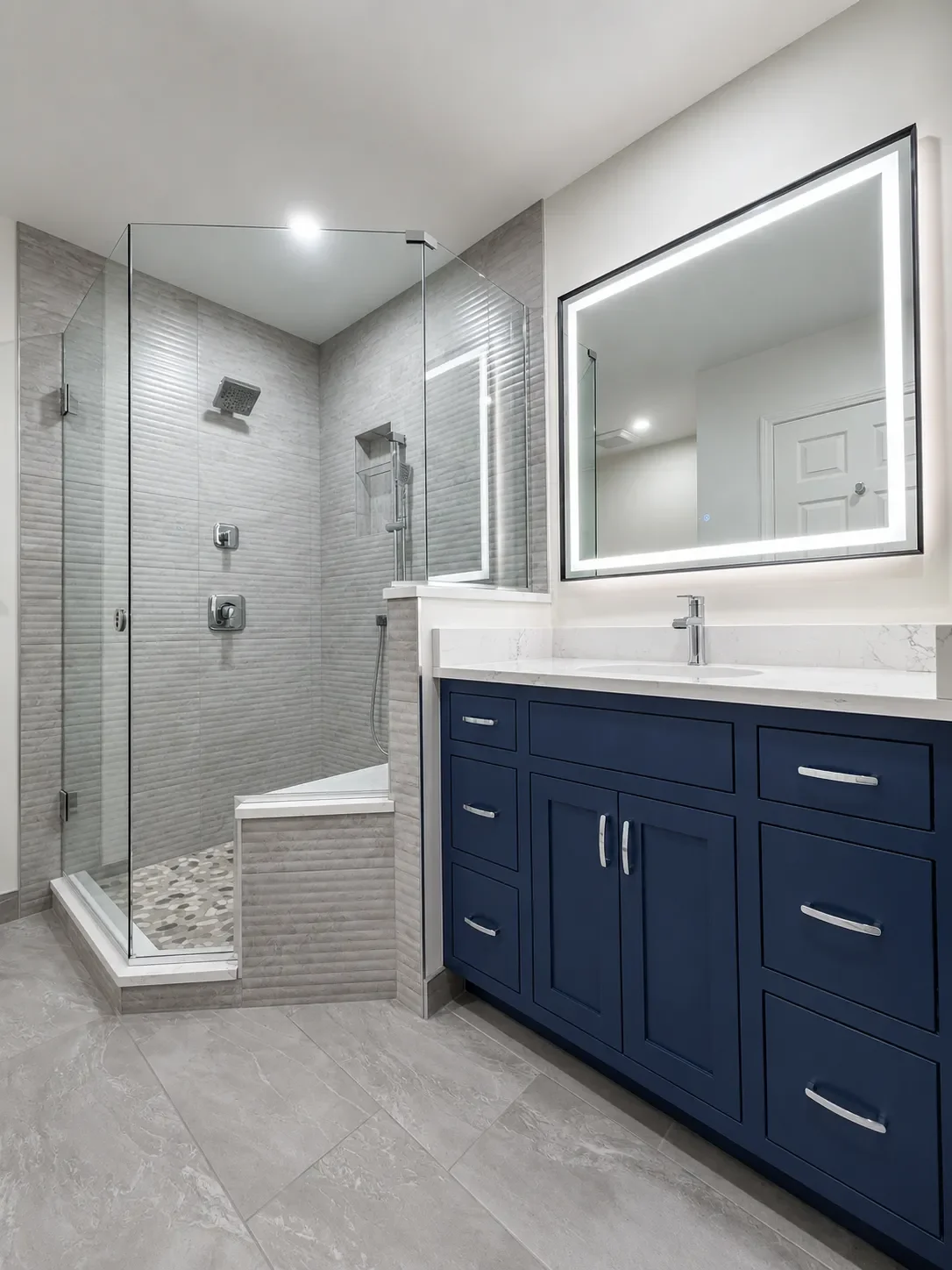 Gunbarrel, CO contemporary master bathroom remodel featuring custom blue vanity, gray stone tile floors, and a glass walk-in shower with gray fluted shower tiles, bench seating, multiple showerheads, and river rock tile floor