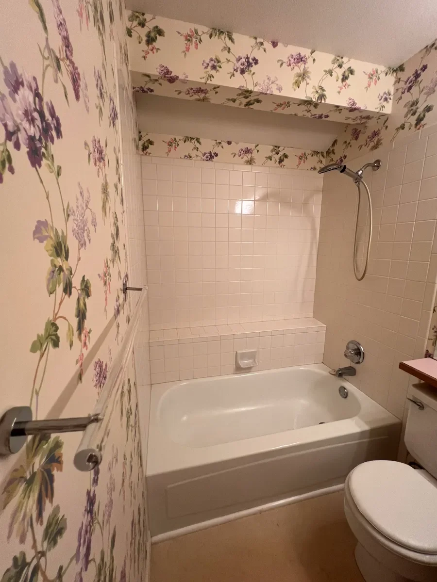 Guest bathroom awaiting renovation, view of shallow white bathtub, A-frame tub overhang, protruding full width tile shelf, and vintage floral wallpaper in Gunbarrel, CO
