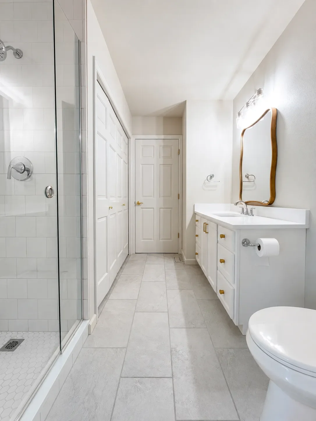 Long portrait view of minimal white contemporary bathroom remodel with walk-in shower, large panel stone tile floors, white vanity with gold hardware, and laundry closet in Gunbarrel, CO