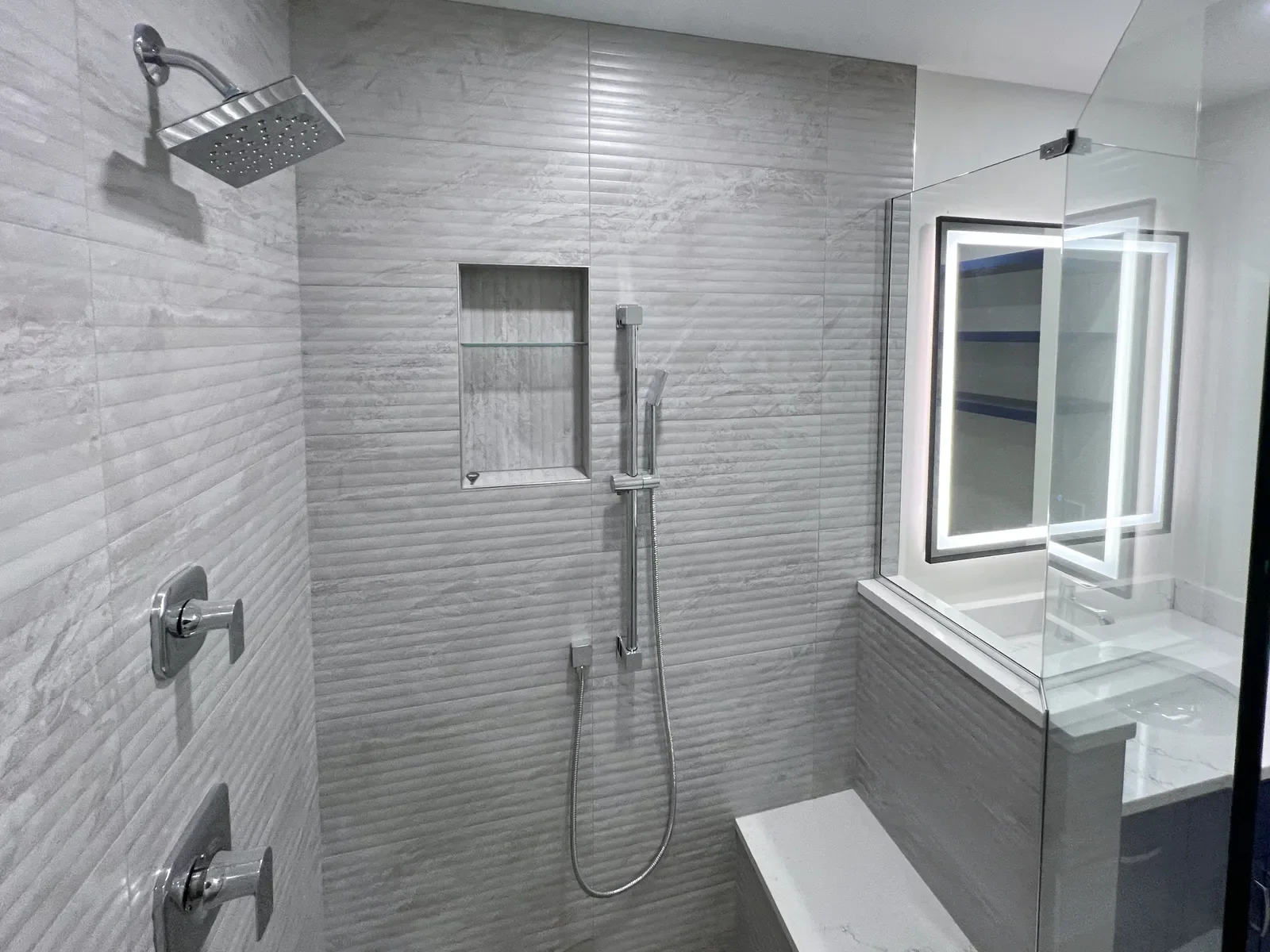 Completed master bathroom remodel in Gunbarrel, CO- view of walk-in shower interior with rain shower head, seated shower head, white quartz bench, recessed niche, and gray textured fluted shower tile