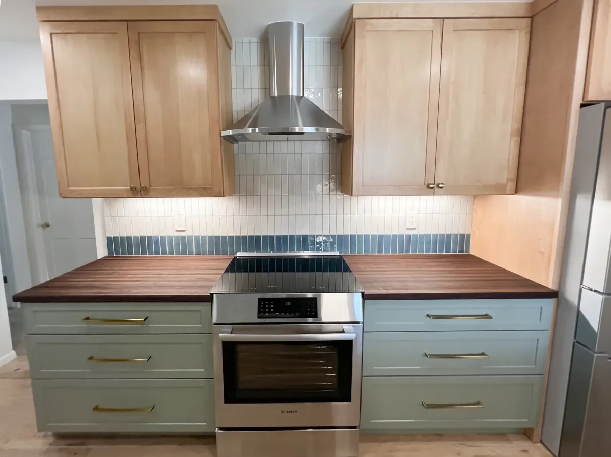 Stove view of remodeled kitchen in Longmont, CO with stainless steel stove and range hood, dark walnut butcher block countertops, maple upper cabinets, sage green lower cabinets, white and teal tile backsplash, and brass drawer pulls