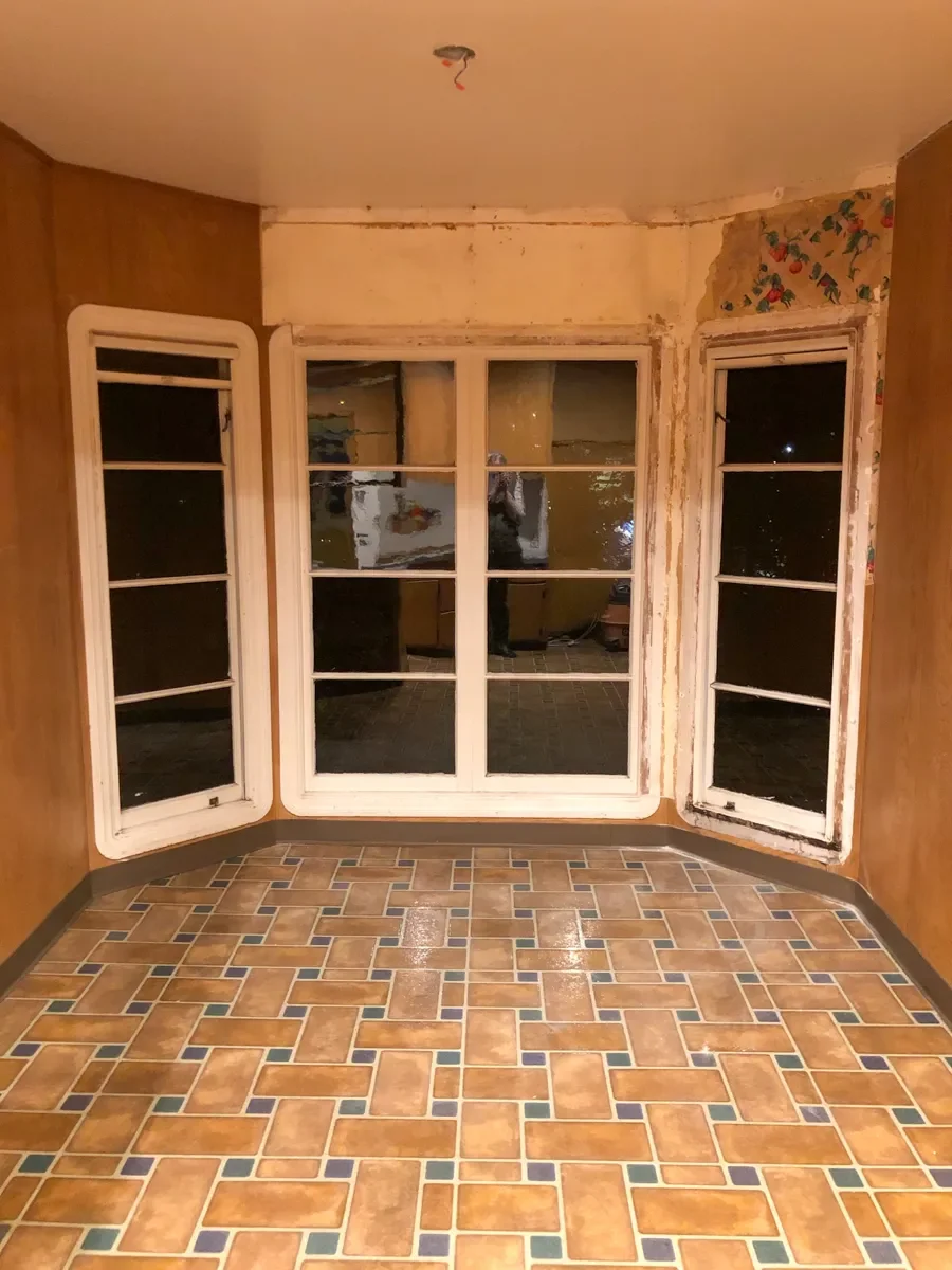 Dining room in Louisville, CO before kitchen remodel featuring floor to ceiling bay windows, peeling vintage wallpaper, and linoleum floors