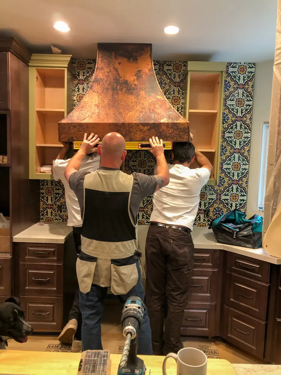 Three workers installing a large copper range hood using a level in a partially completed Tuscan Mediterranean kitchen remodel in Louisville, CO