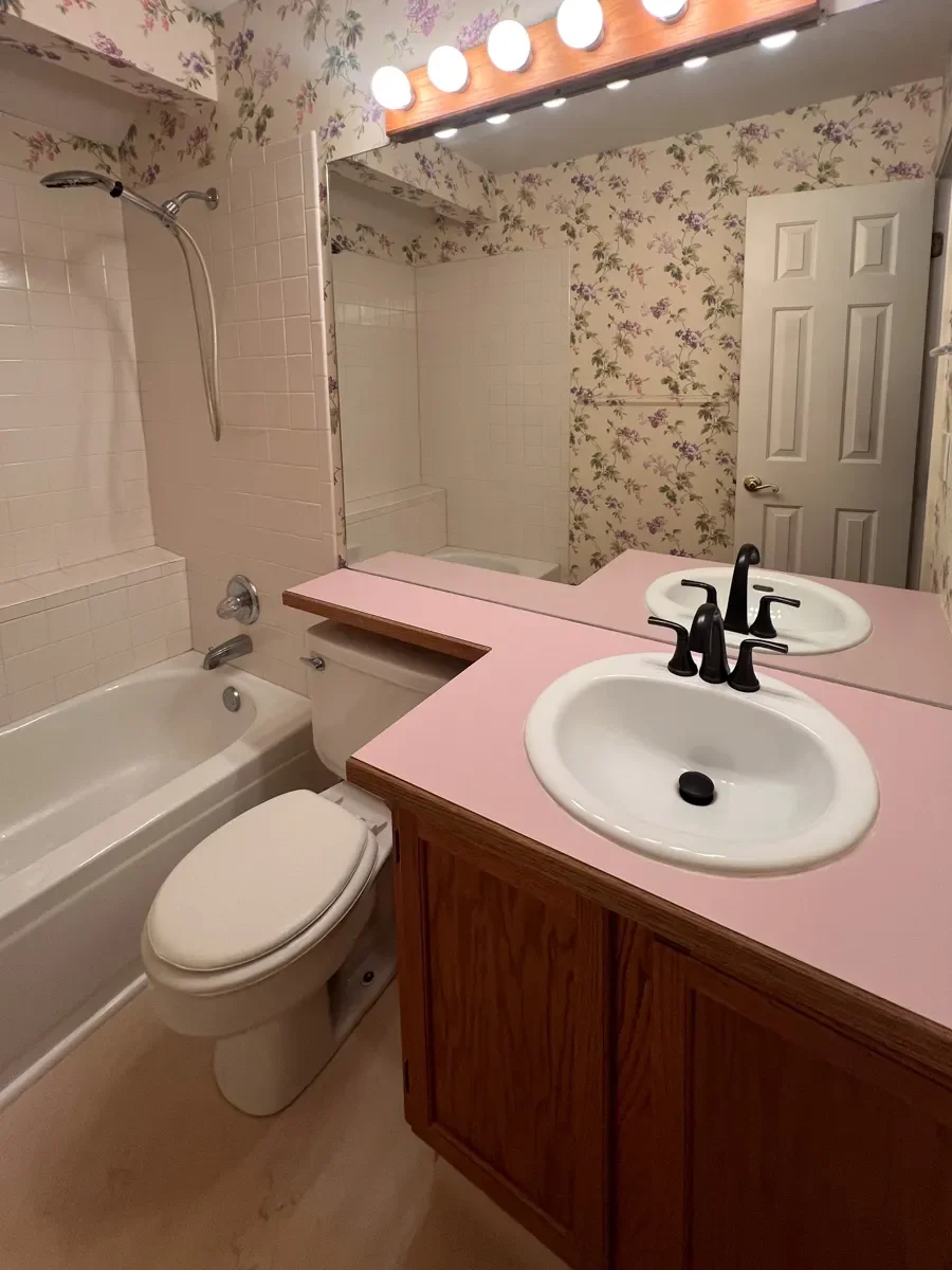 View of old vanity in a Gunbarrel, CO guest bathroom before renovation, with pink laminate countertop, awkward overhang obstructing toilet tank, and outdated wooden vanity with wall-to-wall frameless mirror