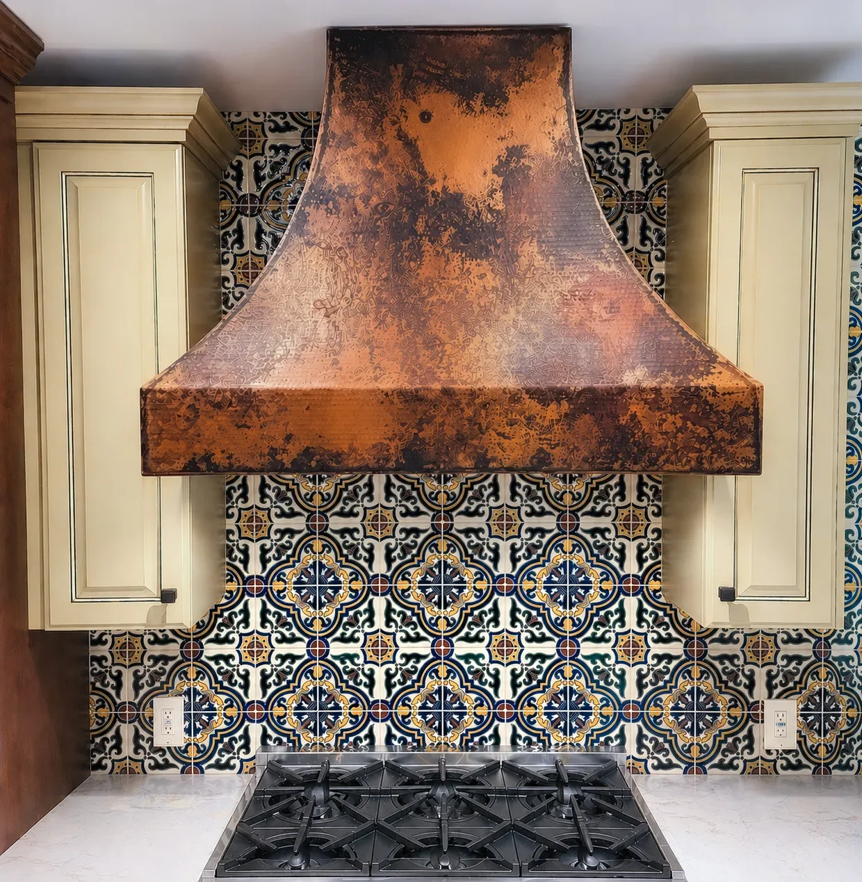 Close up view of spanish patterned tile backsplash, custom yellow crowned cabinets, and copper range hood in an old-world Mediterranean kitchen remodel in Louisville, CO