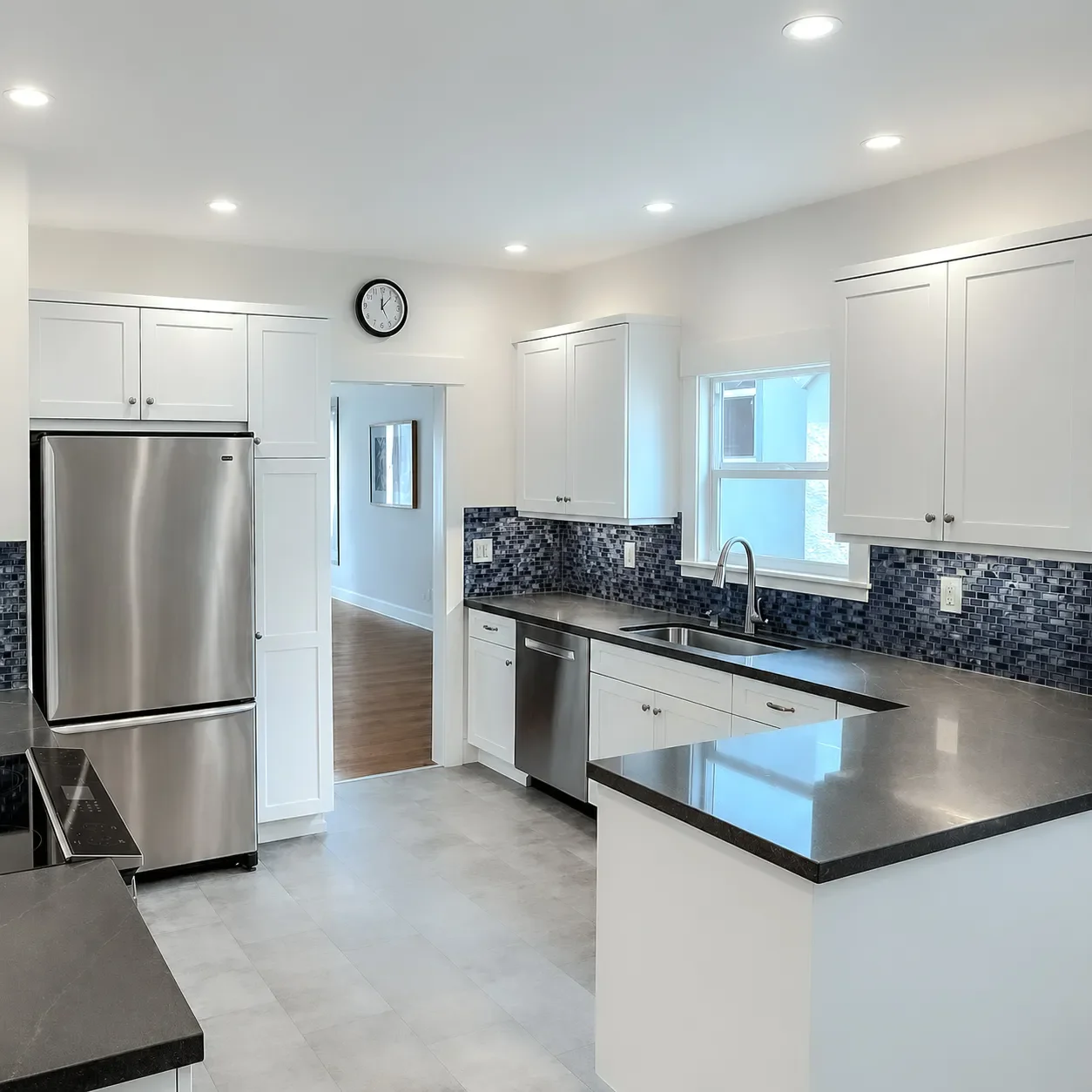 Modern kitchen with stainless steel appliances, white cabinets, black countertops, and gray tile flooring. Features a refrigerator, dishwasher, and a side window, with a wall clock above the doorway.
