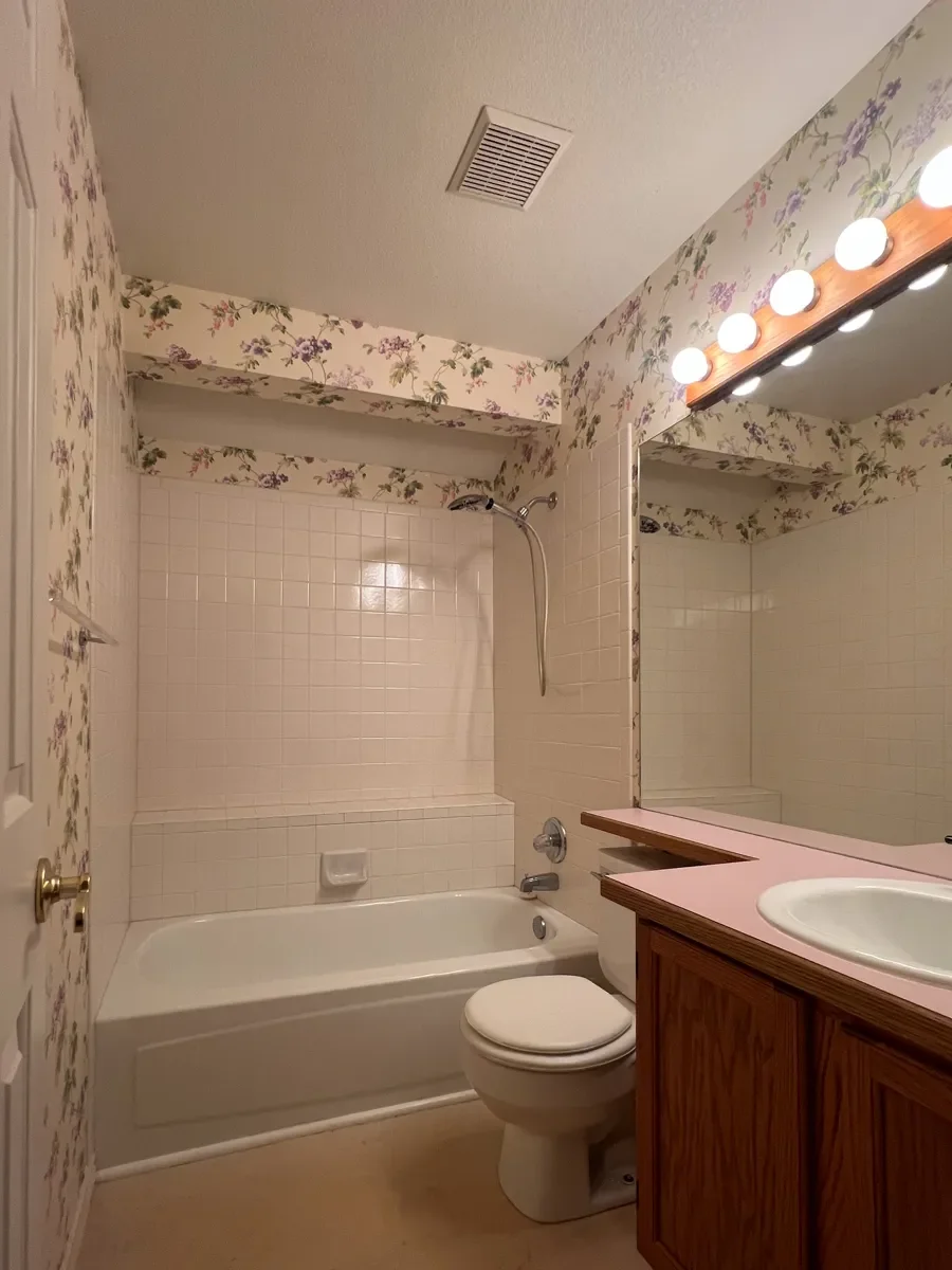 Outdated guest bathroom before renovation showing pink vanity countertop covering toilet tank, linoleum flooring, and vintage floral wallpaper in a Gunbarrel, CO home
