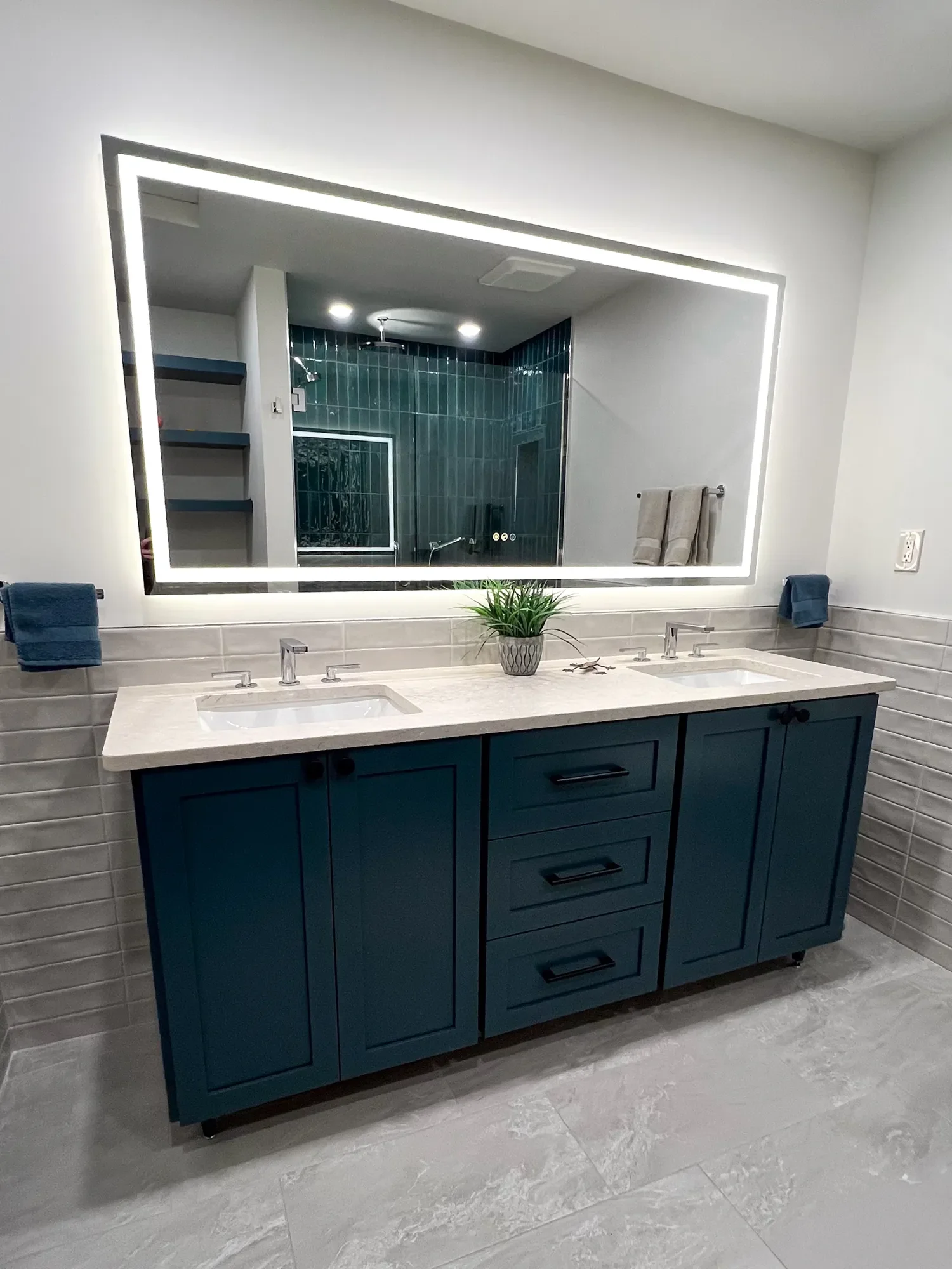 View of custom blue double vanity with large LED mirror reflecting matching built-in floating shelf storage and teal-tiled walk-in shower in a contemporary spa bathroom remodel in the Chautauqua neighborhood of Boulder, CO