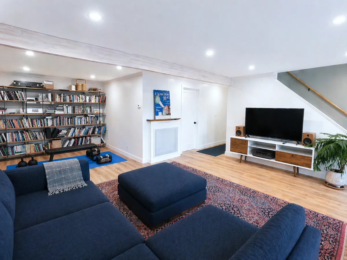 Living room with blue sectional sofa, bookshelf filled with books, large flat-screen TV on a white stand, two speakers, potted plant, decorative rug, dumbbells near a blue mat, and staircase in modern home.