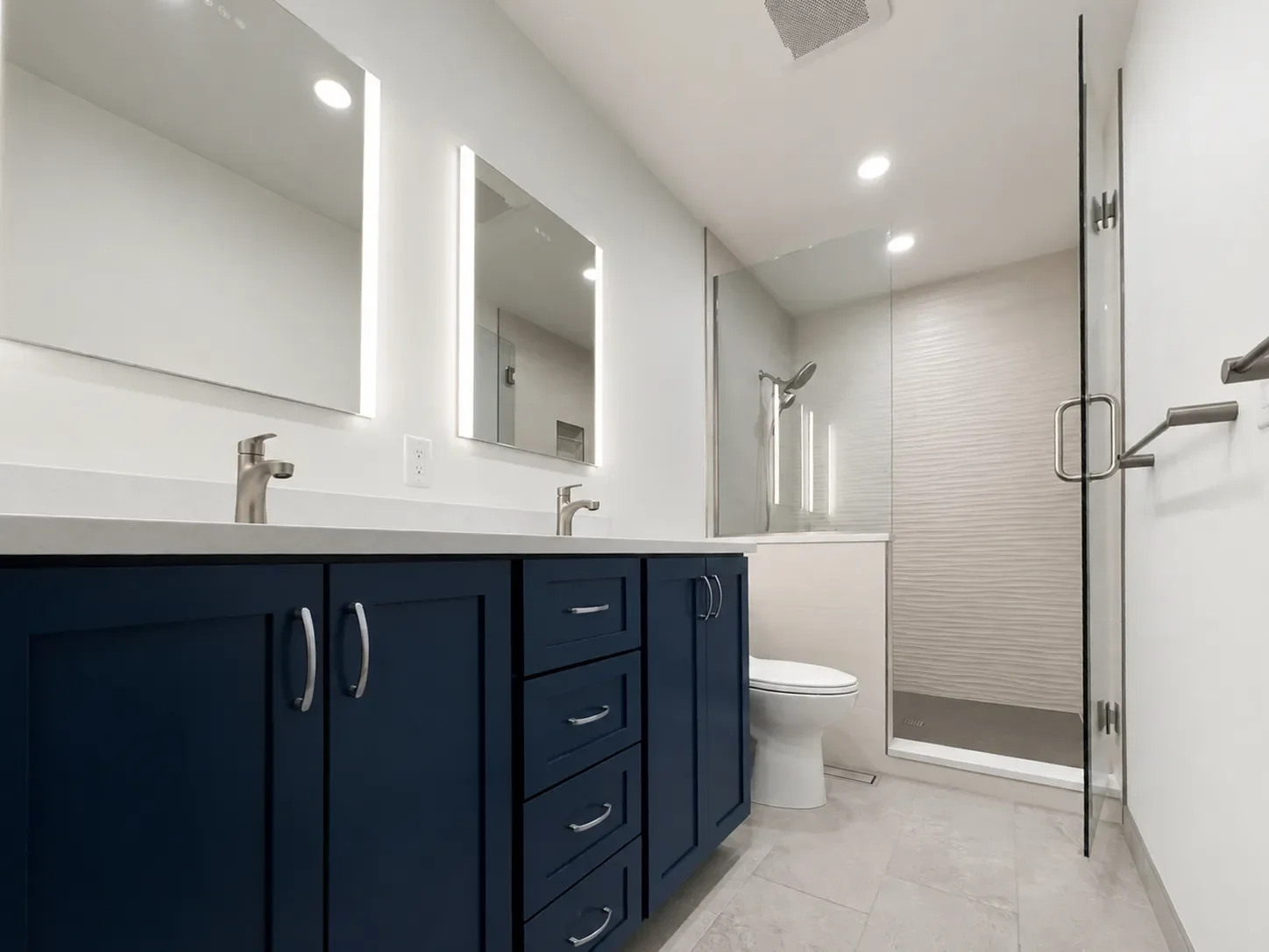Remodeled contemporary bathroom in Boulder, CO with custom navy blue double vanity, recessed medicine cabinets with LED mirrors, frameless walk-in shower with textured accent wall, and gray stone tile floors.