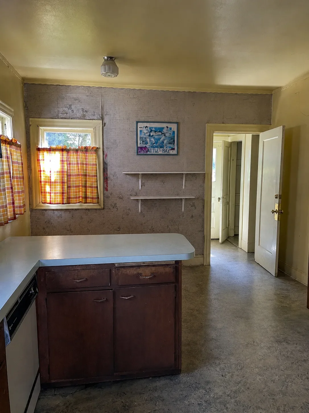 A dark outdated kitchen in desperate need of remodeling in Lafayette, CO featuring peeling brown wallpaper, a tiny ceiling lamp, degraded brown cabinets, linoleum flooring, yellow walls, ceiling, and trim, and powder blue countertops
