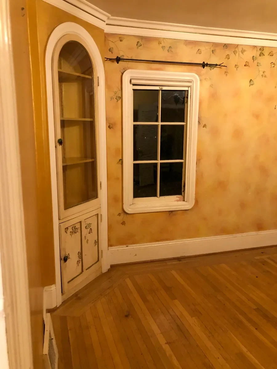 Before photo of kitchen remodel featuring previous living room built-in wall hutch, retro yellow walls, and window with rounded corner trim