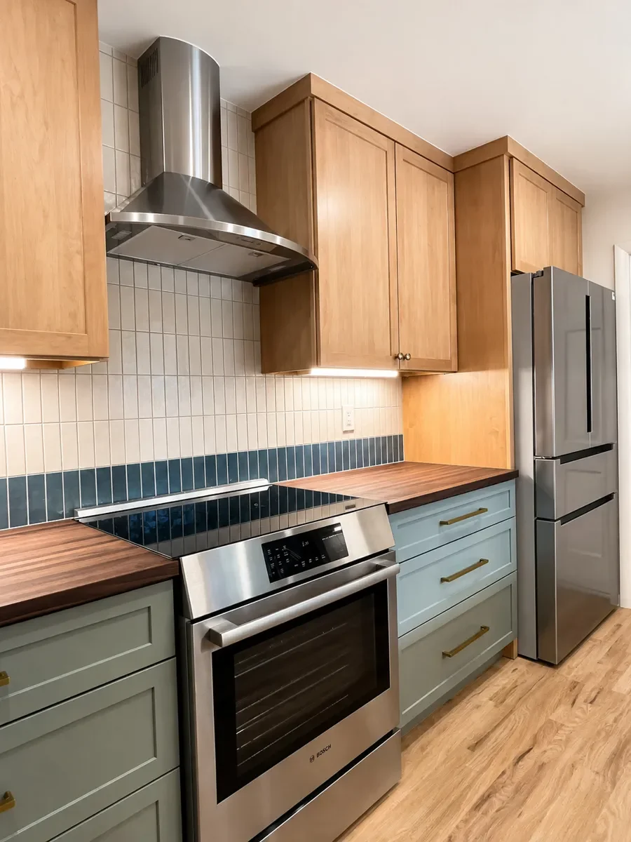 Stove view of remodeled kitchen in Longmont, CO with stainless steel range hood, white and teal full height tile backsplash, dark walnut countertops, maple and sage green cabinets, and red oak hardwood floors.