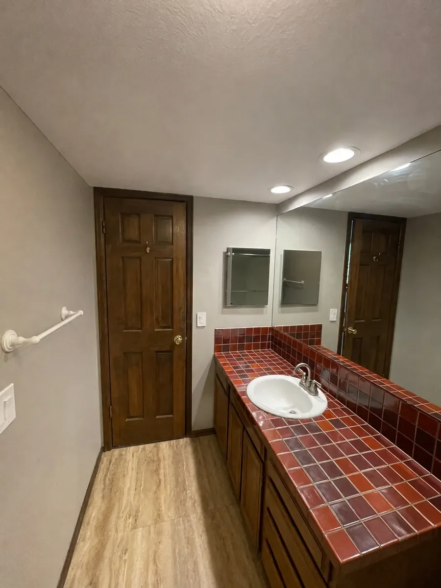 Before pic of Boulder, CO bathroom prior to remodel, featuring a long single vanity with red square tiled countertop, dark wood cabinetry, oversize full wall frameless glass mirror, mirrored medicine cabinet, and white acrylic towel rack