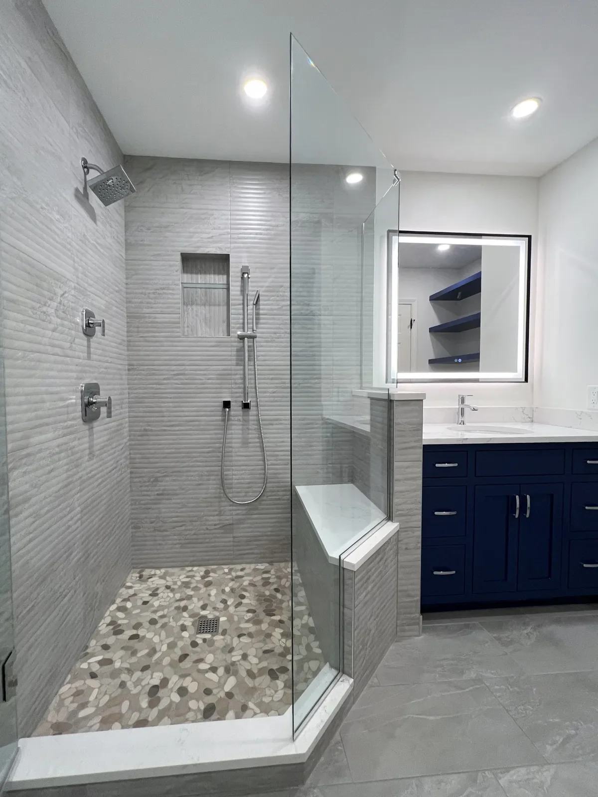 Newly remodeled Gunbarrel, CO master bathroom, neutral minimal contemporary style, featuring walk-in frameless glass shower, textured gray shower tile, large panel gray tile floors, and custom navy blue vanity with dimmable LED mirror