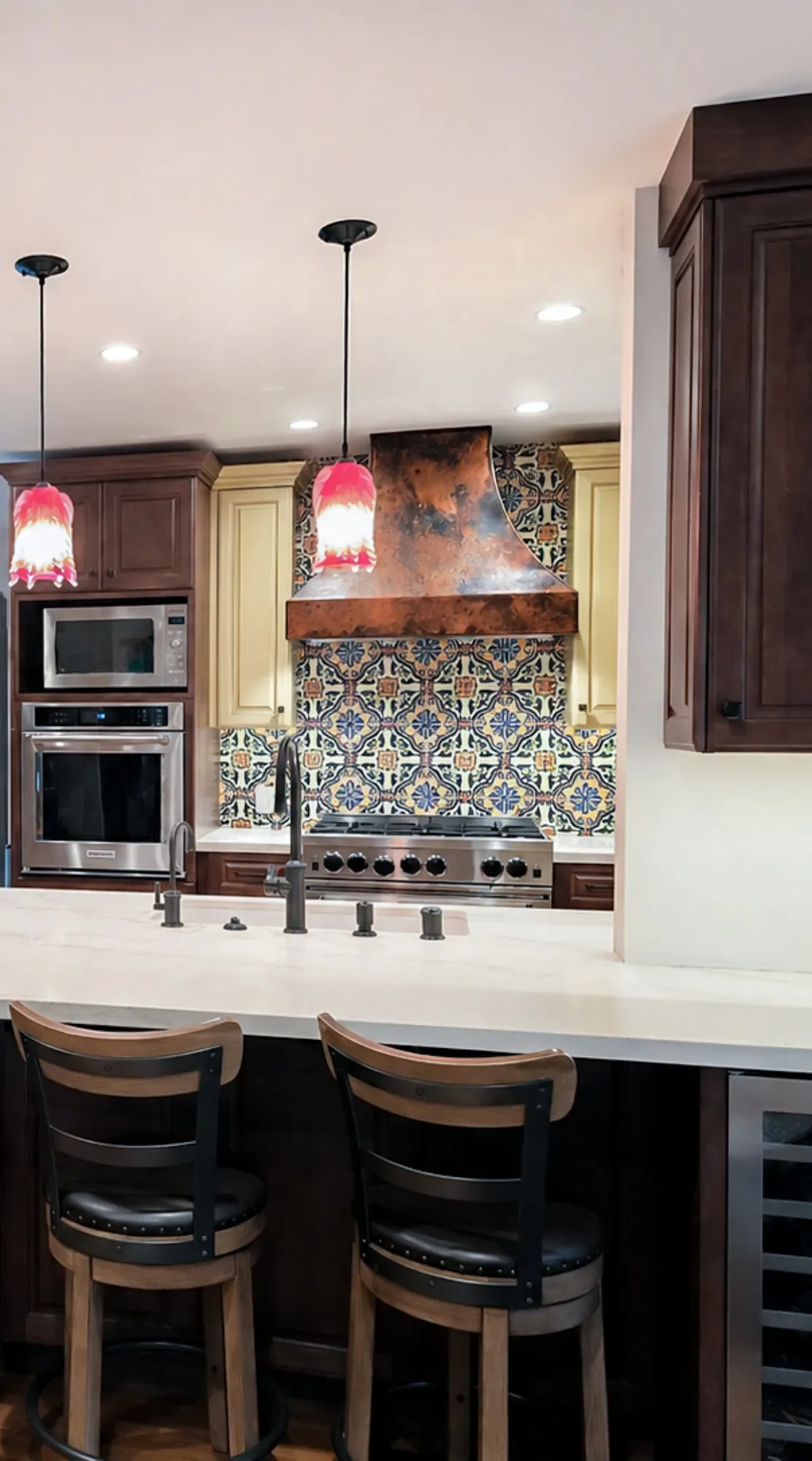 Old-world Mediterranean kitchen remodel in Louisville, CO, featuring white quartz countertops with barstool seating, red blown glass pendant lights, pattern tile backsplash, copper stove range hood, and two-tone cabinets
