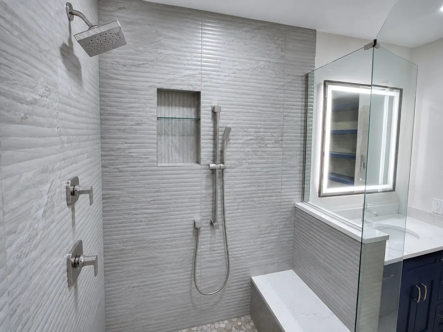 Contemporary master bathroom remodel in Gunbarrel, CO featuring large custom frameless walk-in shower with quartz top bench, storage niche, rain shower head, seated showerhead, and gray fluted tile, adjacent to a custom blue vanity with LED mirror
