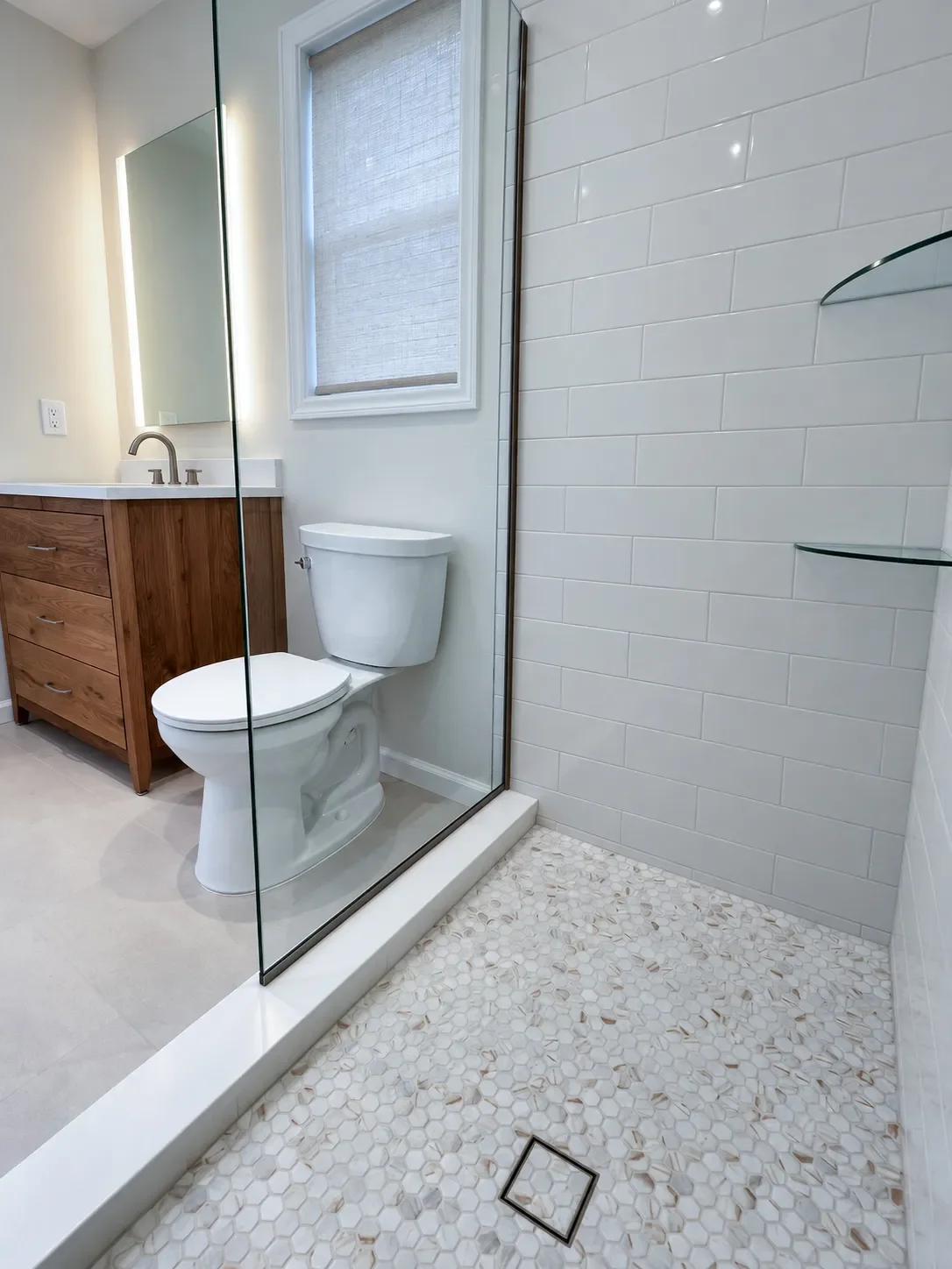 White hexagon mosaic shower tile floor, sleek square tile insert shower drain, white subway wall tile, and glass corner shelves in a frameless glass walk-in shower inside a newly remodeled Boulder, CO bathroom by Downy Home Remodeling