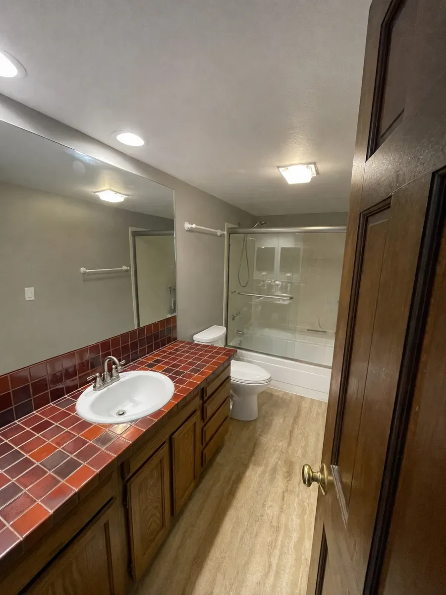  Boulder, CO bathroom awaiting remodel with red tiled dark wood vanity countertop, single oval sink, oversize frameless full height mirror, low ceiling, and sliding glass bathtub shower combo with full length protruding wall shelf