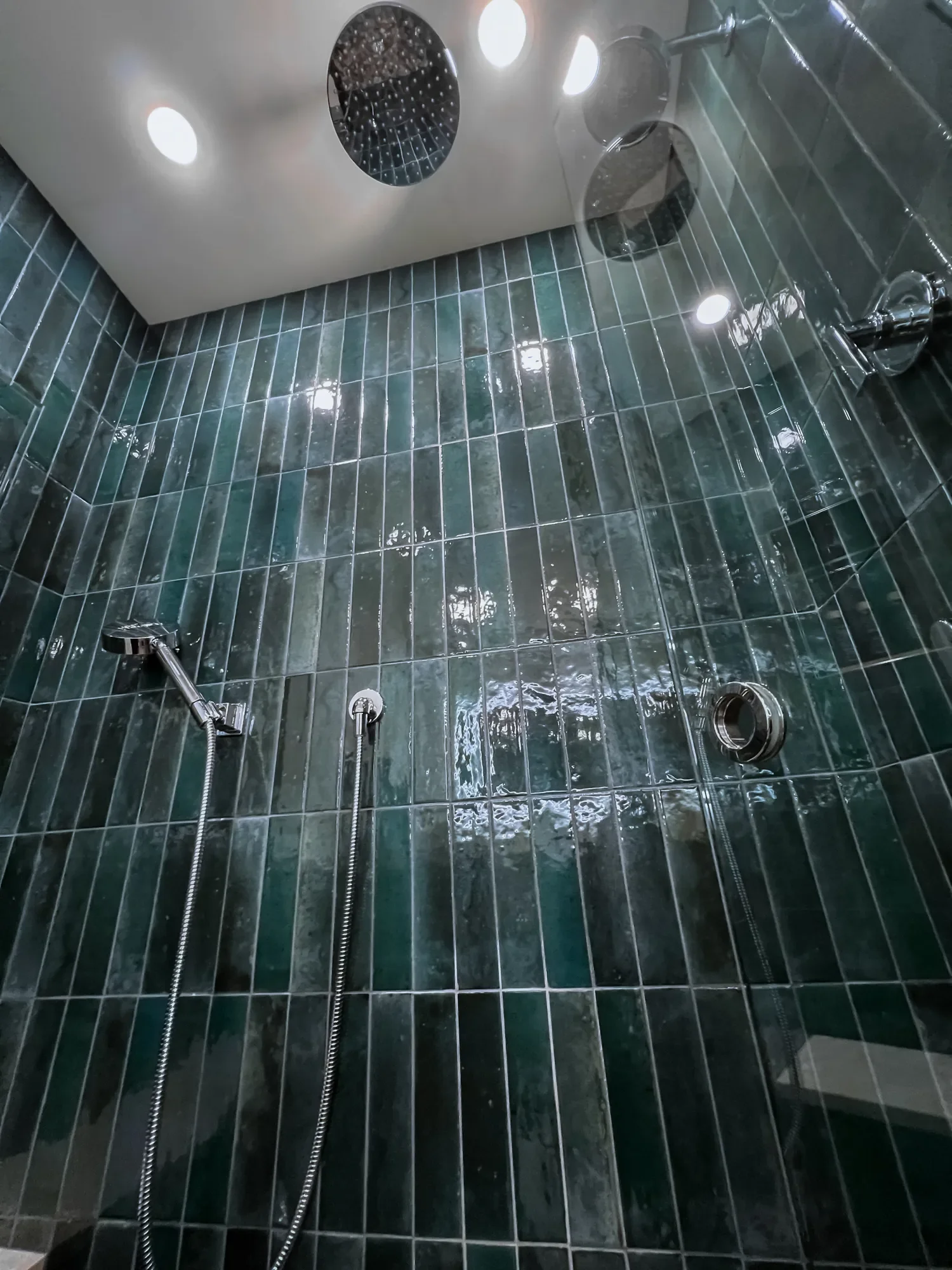 Close-up detail view of a teal tiled walk-in shower interior featuring swinging glass door with silver hole handle, ceiling-mounted rainshower, seated showerhead, and regular wall-mounted showerhead in a Boulder, CO bathroom remodel