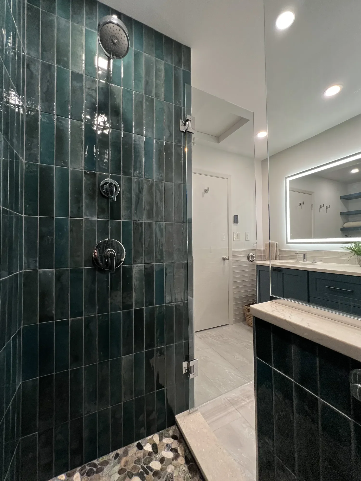 View from teal tiled walk-in shower interior of completed contemporary bathroom remodel in Chautauqua, Boulder, CO looking out at heated stone tiled floor, beige tile wainscoting, custom blue vanity, and extra cabinet storage with floating shelves