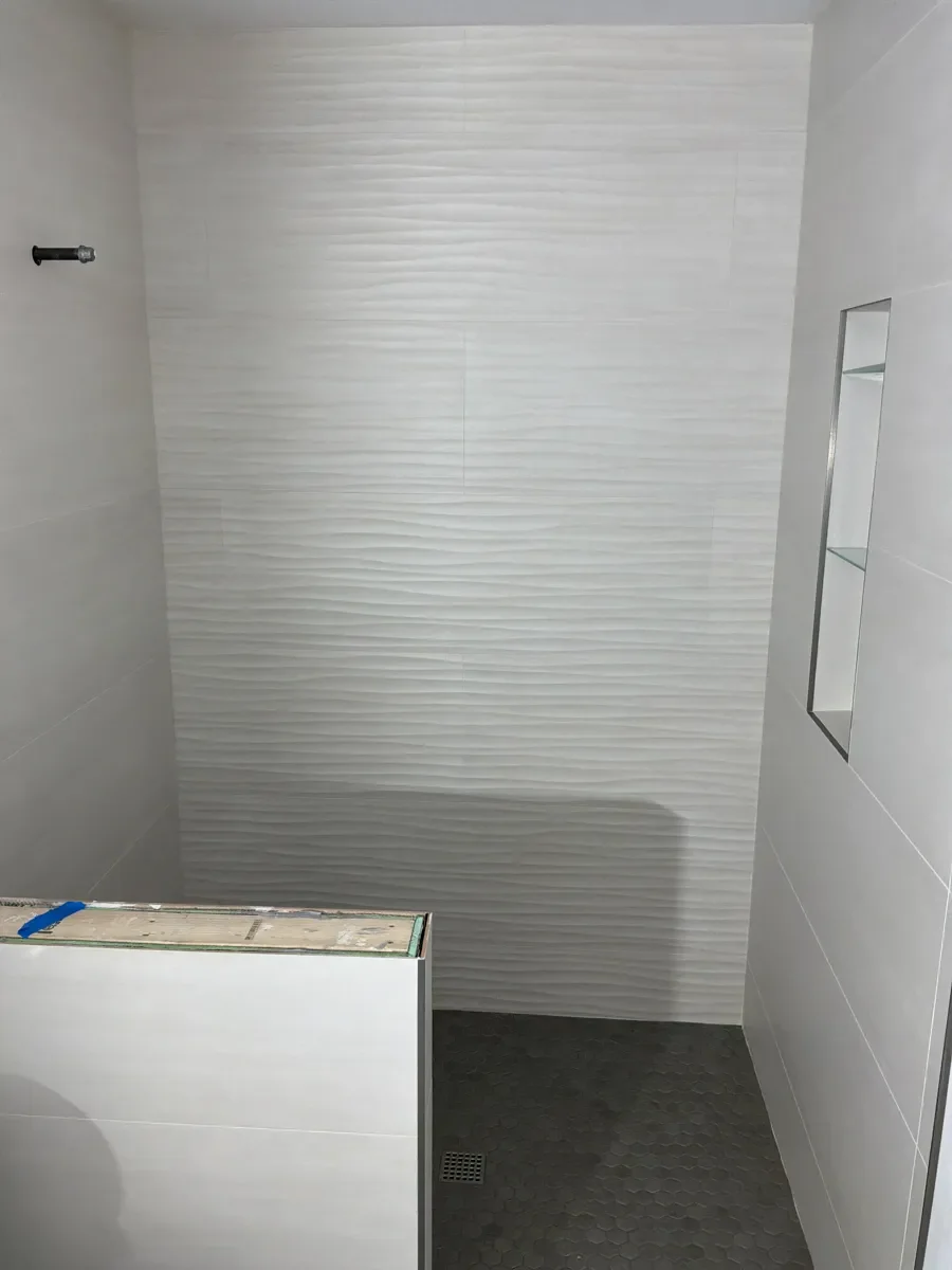 Unfinished bathroom remodel under construction in Table Mesa Boulder, CO featuring newly set large panel textured shower tile, recessed niche with glass shelves, and exposed plumbing awaiting fixtures