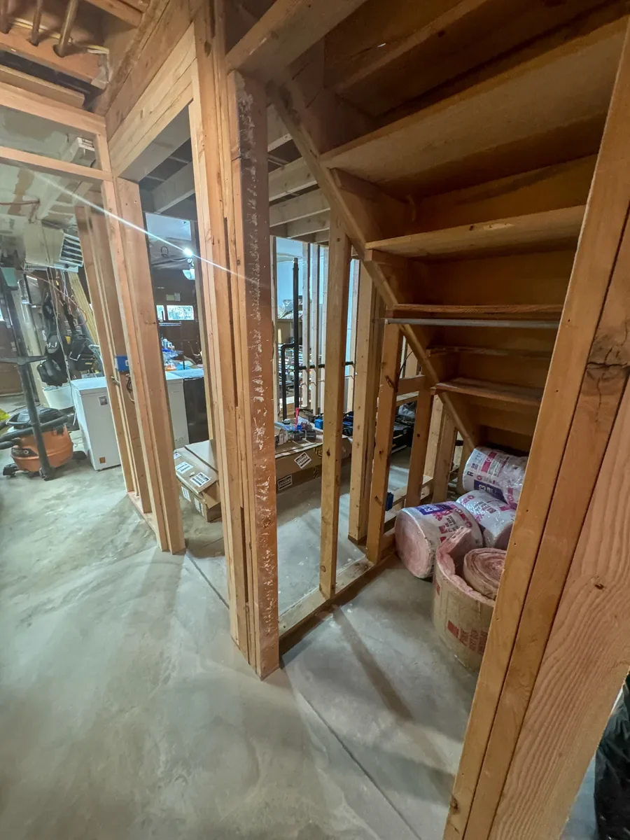 Unfinished basement remodel under construction with exposed wooden framing, unfinished stairs, insulation rolls, and scattered tools on concrete floor