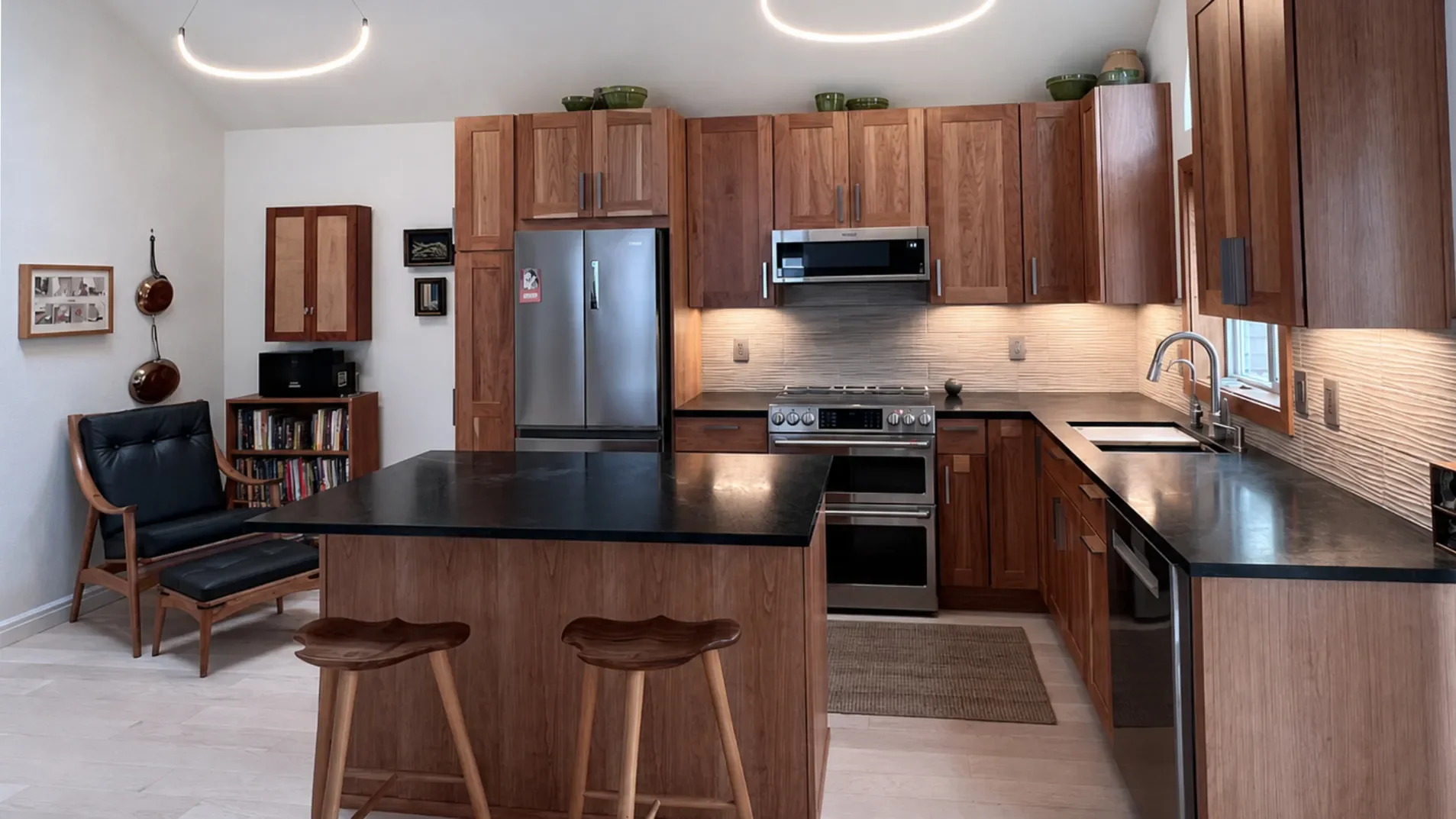 Banner view of completed contemporary craftsman kitchen remodel in North Boulder, CO featuring custom cherry wood cabinets, textured ivory tile backsplash, undercabinet LED lighting, island barstool seating, and adjacent reading nook