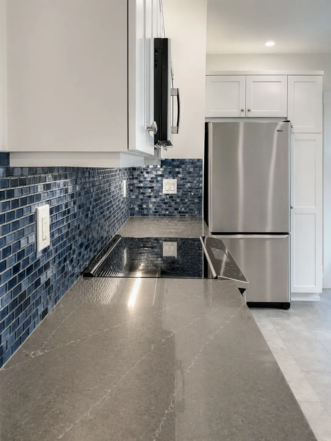 Modern kitchen interior with gray countertops, blue mosaic tile backsplash, stainless steel refrigerator, and white cabinets.