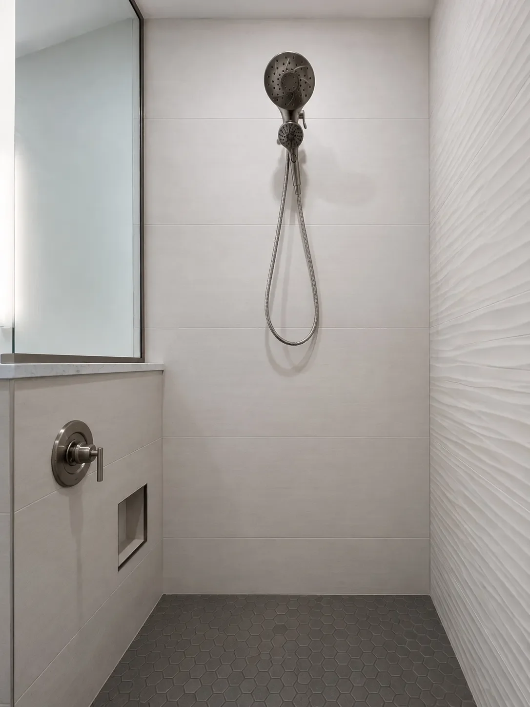 Glass walk-in shower view of bathroom remodel in Table Mesa, Boulder, CO with smooth ivory tile, wavy textured ivory tile accent wall, silver schluter, shaving foot niche, and brushed nickel showerhead with dettachable hand sprayer