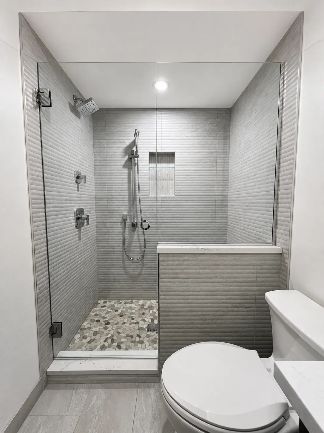 View of walk-in shower in completed guest bathroom remodel in Gunbarrel, CO with gray textured shower tile, rainshower head, seated showerhead, recessed niche with glass shelf, pebble mosaic floor tile, and frameless glass enclosure