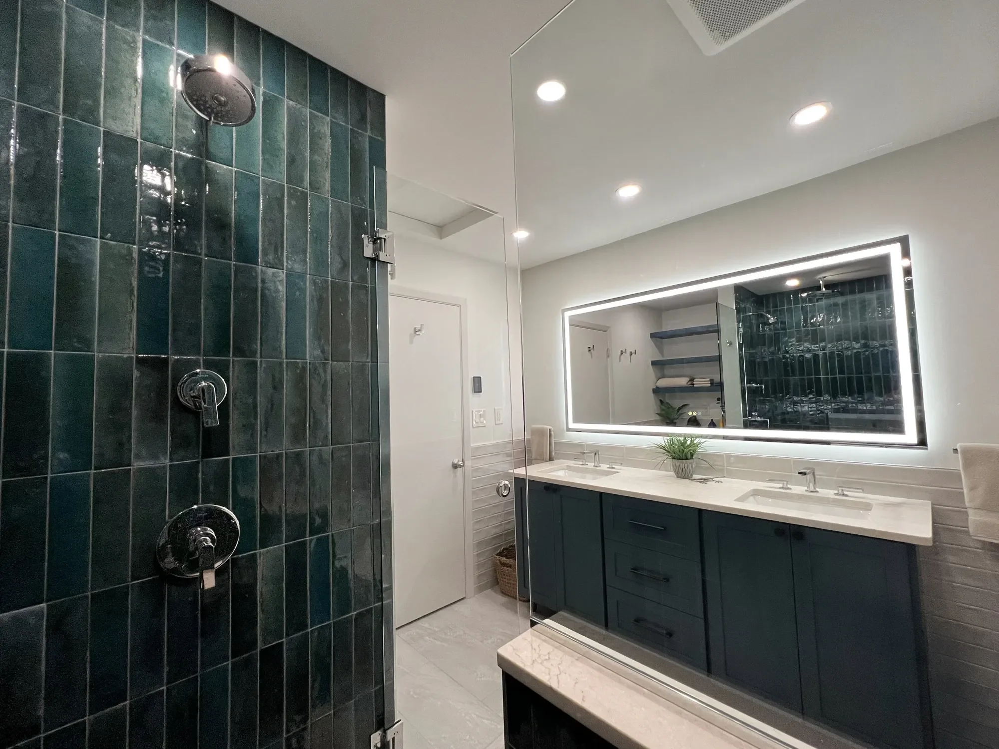 View from walk-in swinging glass door shower showcasing teal soldiered shower tile, custom quartz top double vanity with large LED mirror, beige tile wainscoting and built-in floating shelf storage in a Chautauqua, Boulder, CO bathroom remodel