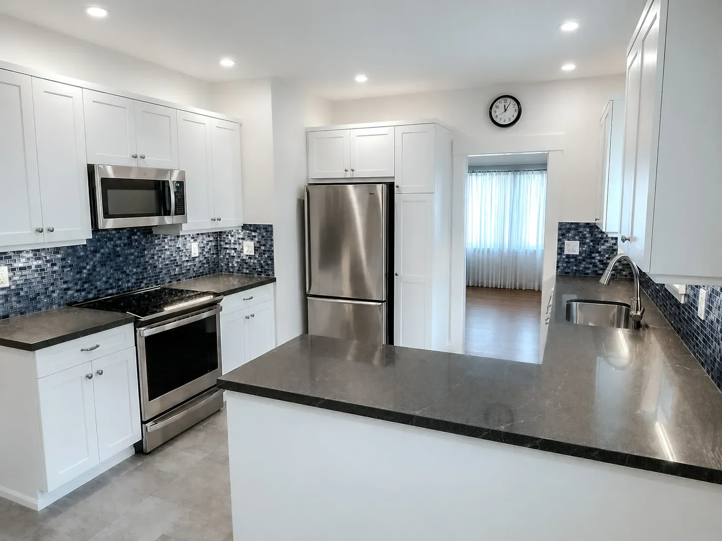 Modern kitchen with white cabinets, stainless steel appliances, dark countertops, and a blue mosaic tile backsplash. A clock is on the wall above the doorway leading to a dining area.