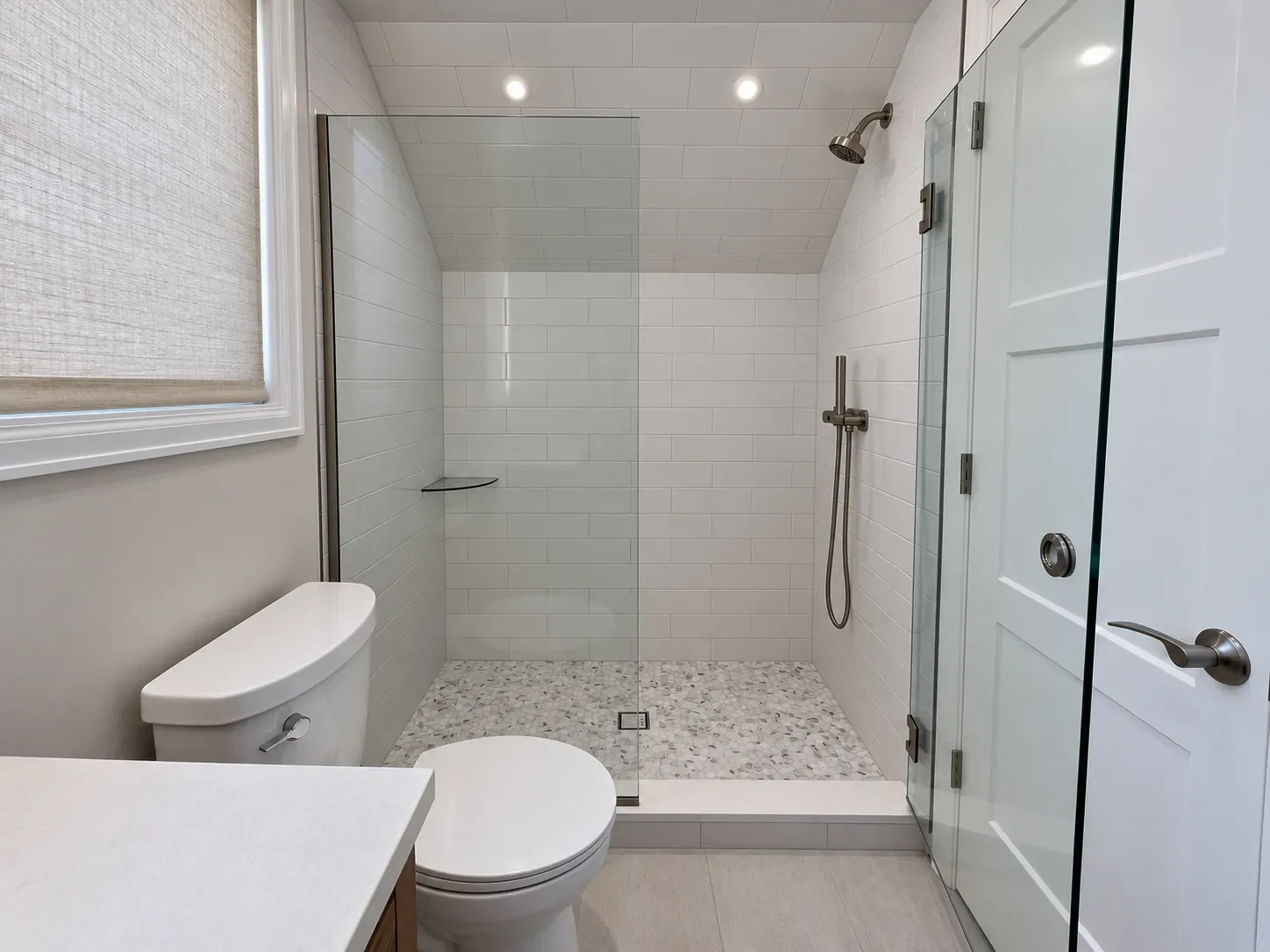 Minimal contemporary style bathroom, newly remodeled by Downy Home Remodeling in Boulder, CO featuring frameless glass swinging door walk-in shower with handheld showerhead, glass corner wall shelves, and white hexagon mosaic shower floor tile