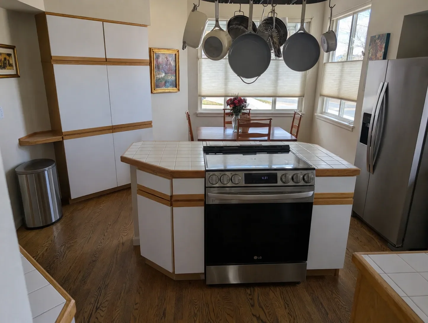 Kitchen awaiting remodel in Gunbarrel, CO with white tile countertops, white laminate cabinets with honey wood trim, and central island housing stainless steel stove with hanging pot rack overhead