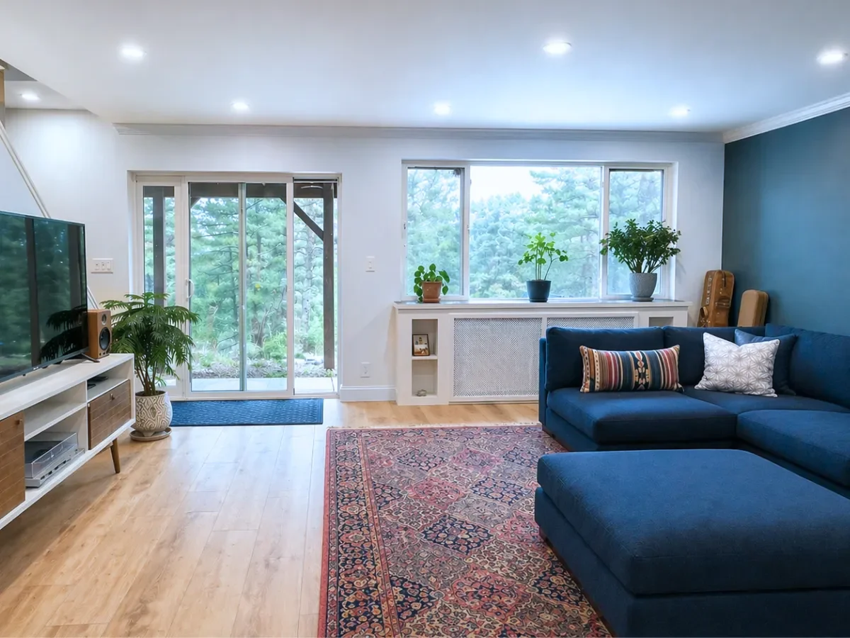 Living room with a blue sectional sofa, large TV, patterned rug, sliding glass doors, and indoor plants. The room has wooden flooring and a blue accent wall.