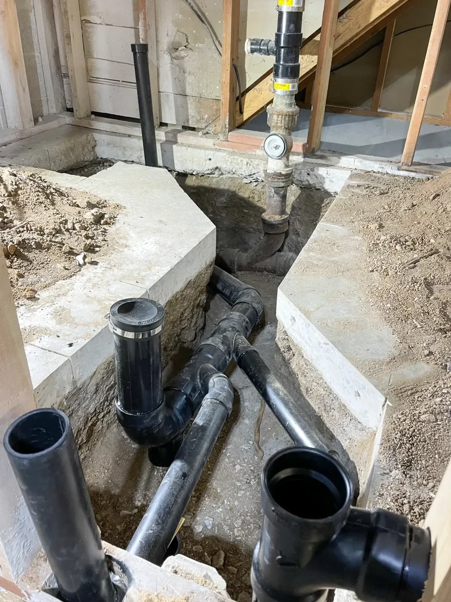 Basement remodel plumbing construction with black PVC pipes exposed in a concrete floor trench with vertical pipe pressure gauge in Boulder, CO